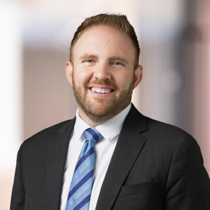Man with short hair and beard wearing a black suit, white shirt, and blue striped tie, stands in front of a blurred indoor background typical of law offices for Chicago lawyers.