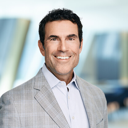 Smiling man with dark hair wearing a light gray plaid suit jacket and white shirt, standing in a modern, blurred corporate law office—a professional among Chicago lawyers.