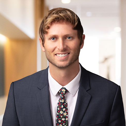 A man in a dark suit and floral tie smiles at the camera, standing in a brightly lit corporate law office hallway.