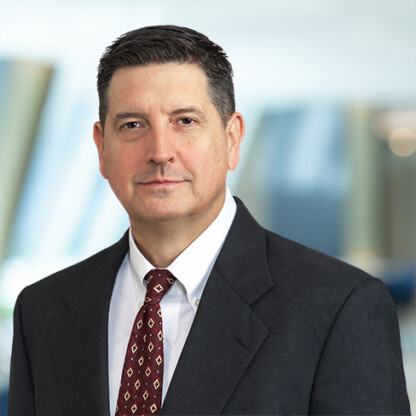 A man in a dark suit, white shirt, and patterned tie stands in front of a blurred corporate law office background, looking at the camera with a neutral expression.