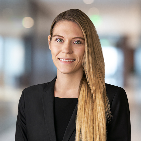 A woman with long straight blonde hair, wearing a black blazer, stands indoors in a blurred law offices setting, facing the camera and smiling slightly.