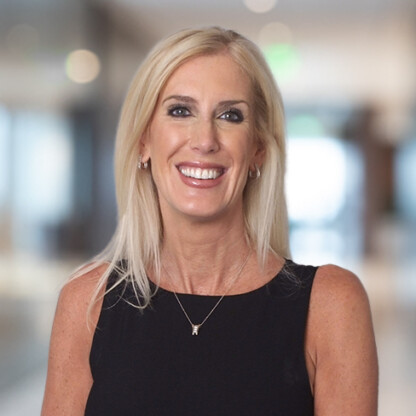 A woman with long blonde hair, wearing a sleeveless black top and a necklace, smiles while standing in a bright, blurred corporate law office setting.