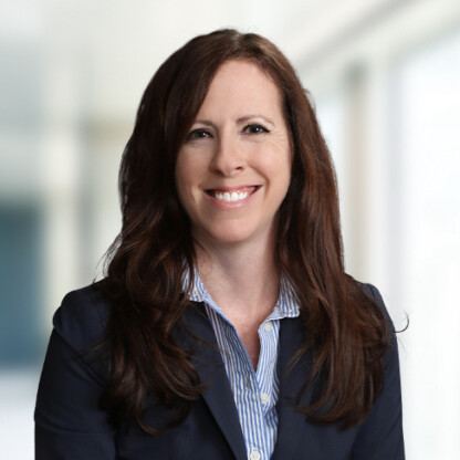 Woman with long brown hair wearing a navy blazer and striped shirt, smiling at the camera in a bright, blurred corporate law office setting.