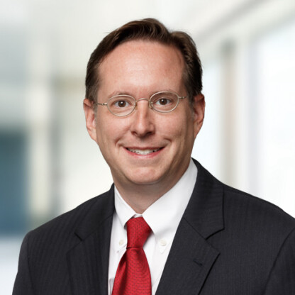 A man wearing glasses, a dark suit, white shirt, and red tie, smiling at the camera in a bright, blurred indoor setting, reflecting the professionalism often seen in law offices specializing in intellectual property law.