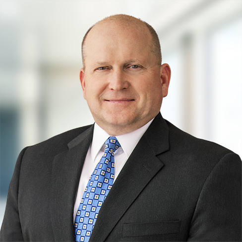 A middle-aged man in a dark suit, white shirt, and patterned blue tie stands indoors, facing the camera with a neutral expression—reflecting the professionalism of lawyers in Chicago at a leading corporate law office.