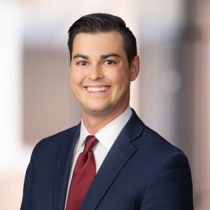A man in a navy suit, white shirt, and red tie smiles at the camera against a blurred corporate law office background, reflecting the professionalism of Chicago lawyers.