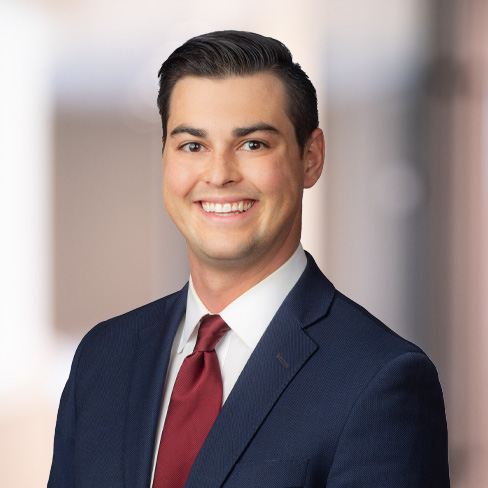 A man in a navy suit, white shirt, and red tie smiles at the camera against a blurred corporate law office background, reflecting the professionalism of Chicago lawyers.