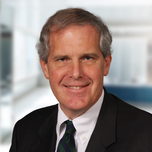 A middle-aged man with gray hair, wearing a dark suit, white shirt, and striped tie, poses in a brightly lit corporate law office.