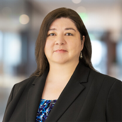 A woman with straight brown hair wearing a black blazer and patterned blouse stands in a modern, blurred corporate law office setting, facing the camera with a neutral expression.