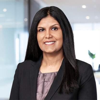 A woman with long dark hair, wearing a dark blazer over a gray blouse, stands in a modern corporate law office with glass walls and bright lighting, reflecting the dynamic environment of Chicago lawyers.