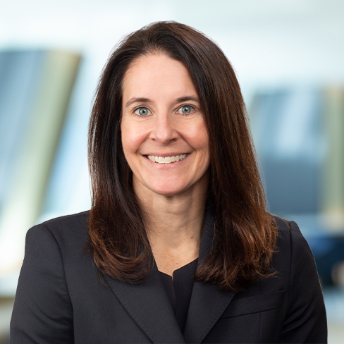 A woman with long brown hair wearing a black blazer smiles at the camera in a professional corporate law office setting.