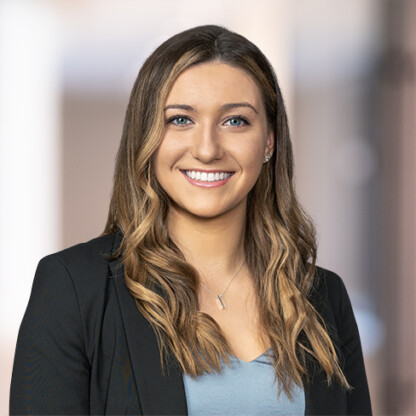 A woman with long wavy brown hair, wearing a black blazer over a light blue top, smiles at the camera in a softly blurred indoor setting, reflecting the professionalism found in leading Chicago lawyers and law offices.