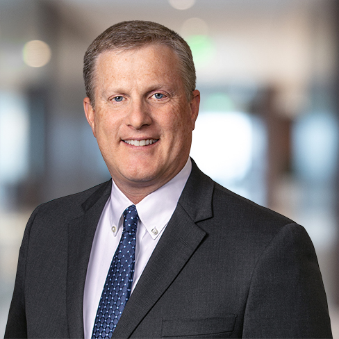 A man in a dark suit and blue tie smiles at the camera in a softly blurred office setting, conveying professionalism often seen in law offices specializing in intellectual property law.