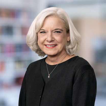 Older woman with short gray hair, wearing a black top and necklace, smiles in front of a blurred indoor background with shelves—reflecting the professionalism of Chicago lawyers experienced in intellectual property law.
