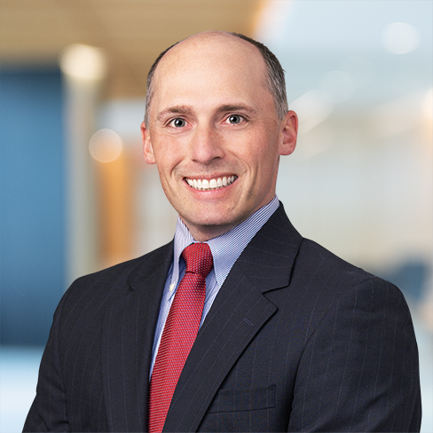 A man in a dark suit, light blue shirt, and red tie smiles at the camera in a modern corporate law office setting.
