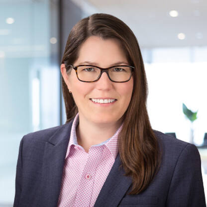 A woman with long brown hair and glasses, wearing a dark blazer and pink patterned shirt, stands in a modern law office setting, smiling at the camera—representing dedicated lawyers in Chicago.