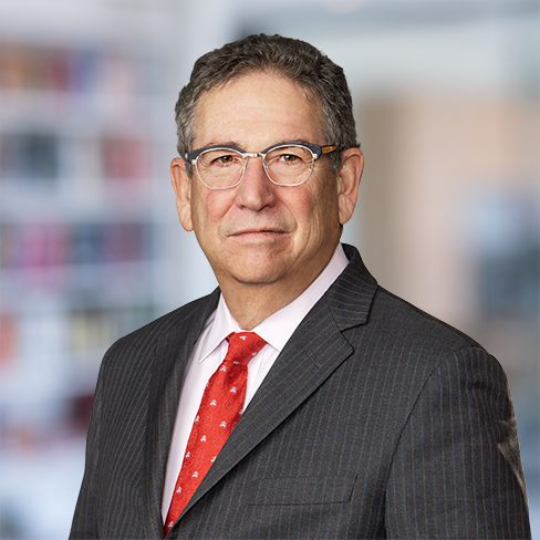 A middle-aged man wearing glasses, a dark pinstripe suit, a white shirt, and a red tie poses in front of a blurred office background, reflecting his expertise in intellectual property law.