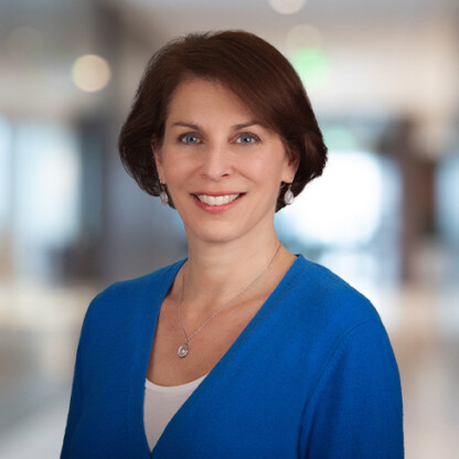 A woman with short brown hair, wearing a blue sweater and a pendant necklace, smiles in a softly blurred indoor setting, embodying professionalism often found in law offices and litigation support environments.