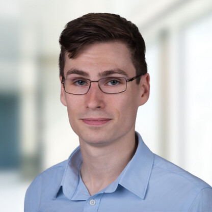 A young man with short brown hair and glasses wearing a light blue collared shirt, standing in a brightly lit corporate law office.