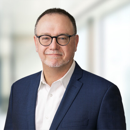 A man wearing glasses, a navy blue suit jacket, and a white shirt stands in front of a blurred indoor background, capturing the professional atmosphere of a corporate law office.