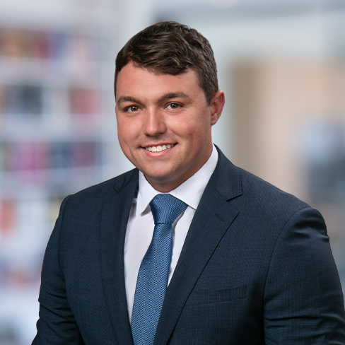 A man in a dark blue suit, white shirt, and blue tie is smiling in front of a blurred office background, reflecting the professionalism of a corporate law office.