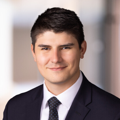 A man wearing a black suit, white shirt, and dotted tie, with short dark hair, poses for a professional headshot against a blurred background—ideal for representing law offices or corporate law office environments.