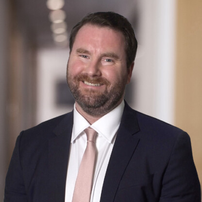 A man with dark hair and a beard, wearing a navy suit, white shirt, and light pink tie, smiles in a blurred corporate law office hallway.