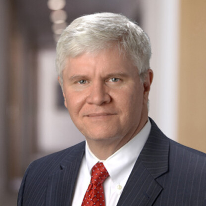 A middle-aged man with short gray hair wearing a dark pinstripe suit, white shirt, and red tie stands confidently in the blurred hallway of a corporate law office.