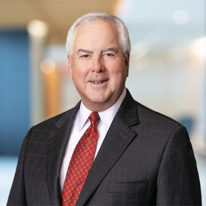 An older man with short white hair wears a dark suit, white shirt, and red patterned tie, standing in a modern, blurred law offices setting.