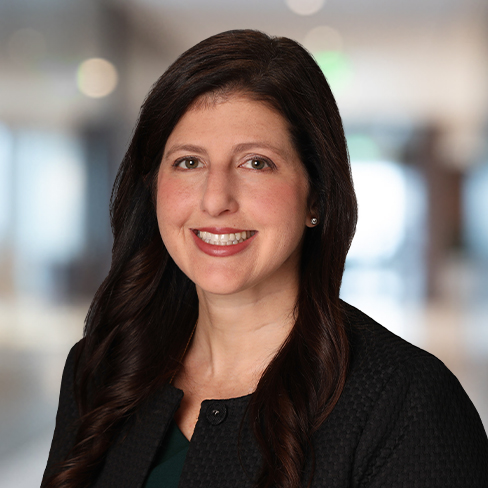 Woman with long dark hair smiles at the camera, wearing a dark textured blazer, in a brightly lit, blurred indoor setting—an inspiring professional among leading lawyers in Chicago.