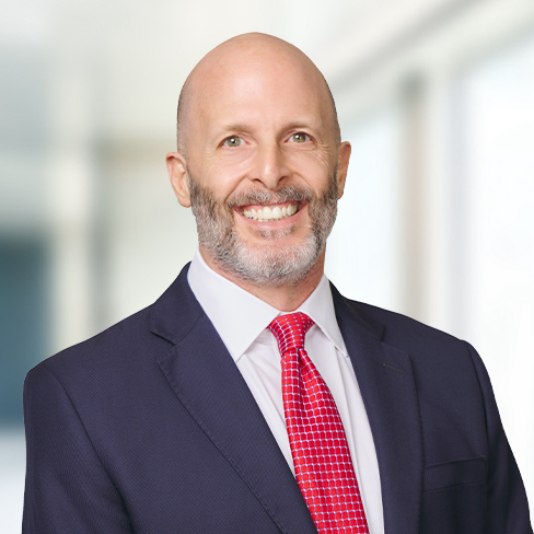 A bald man with a trimmed gray beard, wearing a navy suit, white shirt, and red patterned tie, smiles in a brightly lit law office setting specializing in litigation support.