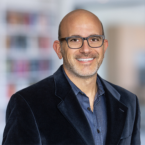 A man with glasses, wearing a dark blazer and blue shirt, smiles at the camera in an indoor setting with blurred shelves in the background of a corporate law office.