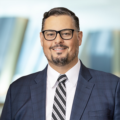 Man wearing glasses, a dark blue suit, white shirt, and striped tie, smiling in front of a blurred background with shelves or books—ideal imagery for corporate law office or litigation support settings.