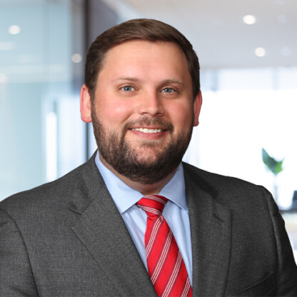 A man in a business suit and red striped tie smiles while standing in a modern corporate law office with glass walls and bright lighting.
