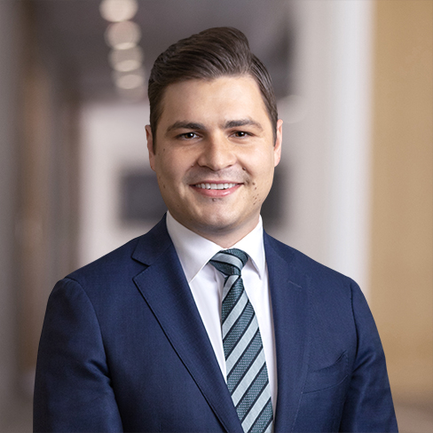A man in a navy blue suit, white shirt, and striped tie smiles while standing in a blurred corporate law office hallway background.