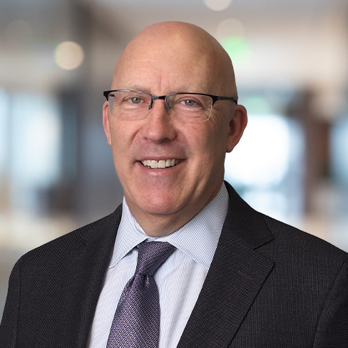 A bald man wearing glasses, a suit jacket, a dress shirt, and a tie, standing indoors with a blurred office background—ideal for profiles of Chicago lawyers specializing in litigation support.
