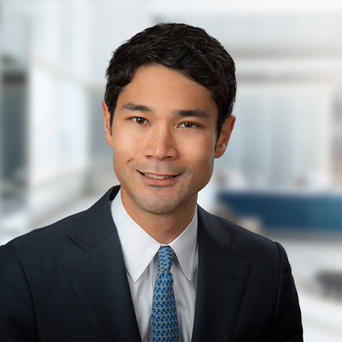 A man in a dark suit and blue tie smiles at the camera against a blurred corporate law office background.