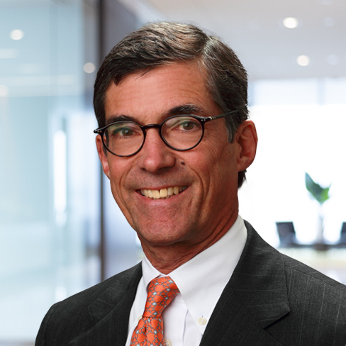 A man wearing glasses, a dark suit, white shirt, and orange patterned tie stands in a modern corporate law office with a blurred background.