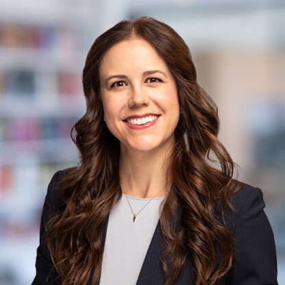 Woman with long brown hair wearing a dark blazer and light top, smiling confidently—an inspiring professional among lawyers in Chicago—set against blurred shelves and warm indoor lighting.