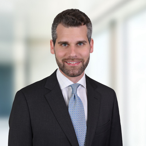 A man in a dark suit, light shirt, and patterned tie stands in front of a blurred corporate law office background, facing the camera and smiling.