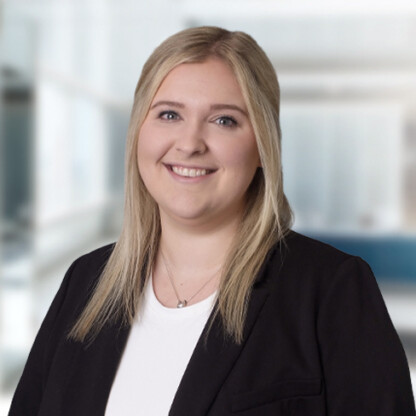 A woman with straight blonde hair, wearing a black blazer over a white top, smiles at the camera in a brightly lit corporate law office setting.