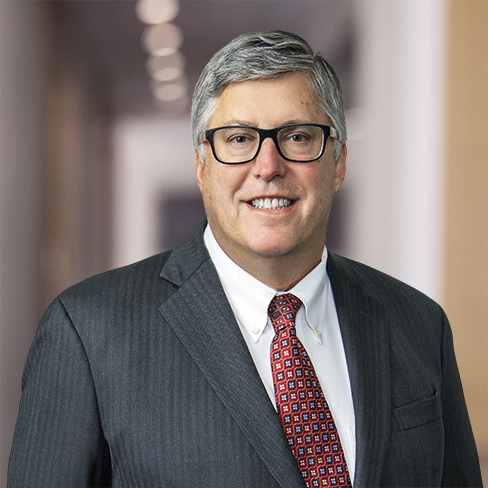 A middle-aged man with gray hair and glasses, wearing a dark suit, white shirt, and red patterned tie, stands in a blurred indoor setting typical of law offices for Chicago lawyers.