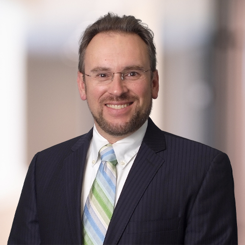 Man in a dark pinstripe suit, white shirt, and striped tie, standing and smiling in front of a blurred indoor background—an embodiment of Chicago lawyers experienced in litigation support.