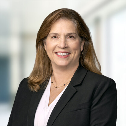 A woman with long brown hair wearing a black blazer, light blouse, and necklace, smiling in a bright, blurred indoor setting—an approachable expert among leading Chicago lawyers.
