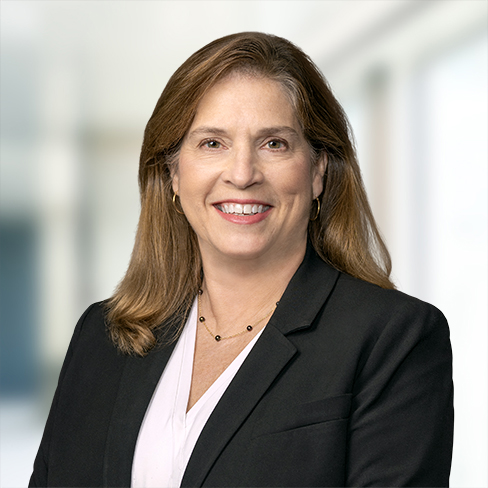 A woman with long brown hair wearing a black blazer, light blouse, and necklace, smiling in a bright, blurred indoor setting—an approachable expert among leading Chicago lawyers.