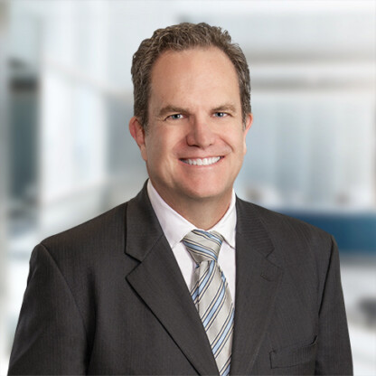 Smiling man in a dark suit, white shirt, and striped tie standing in a bright, modern corporate law office.