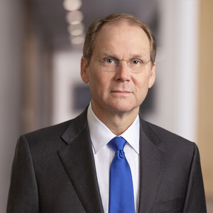 Middle-aged man in a dark suit, white shirt, and blue tie, wearing glasses, standing in a professional indoor setting with blurred background at a corporate law office.
