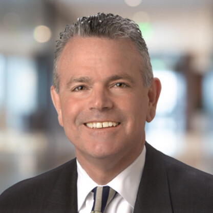 Smiling man in a business suit and tie with short gray hair, posing in a blurred office setting, representing experienced lawyers in Chicago.
