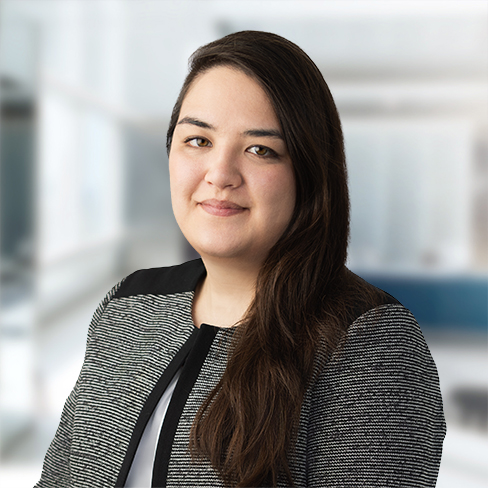A woman with long dark hair wearing a textured gray blazer stands in a modern, bright corporate law office, looking at the camera with a neutral expression.