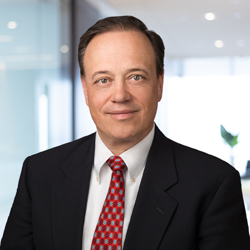 A middle-aged man in a dark suit, white shirt, and red patterned tie poses in a modern corporate law office with a blurred background.